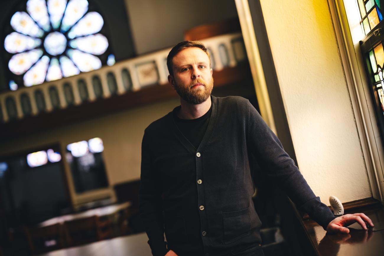Dean Caivano poses for a photo in a church converted into a classroom wearing a dark blue sweater.